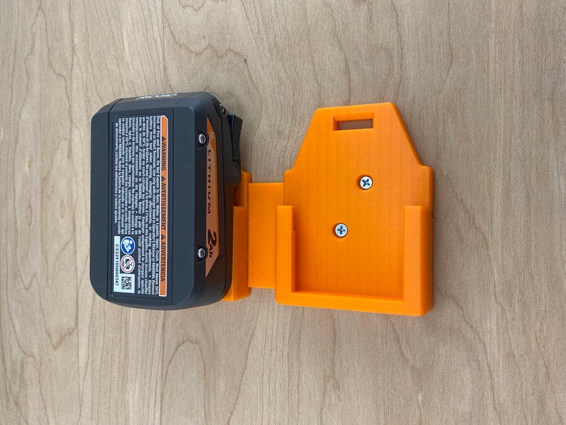 Puede incluir: Un soporte de pared para cargador de bater&iacute;a negro y naranja con dos tornillos. El cargador de bater&iacute;a est&aacute; etiquetado como "2Ah Li-Ion Battery".