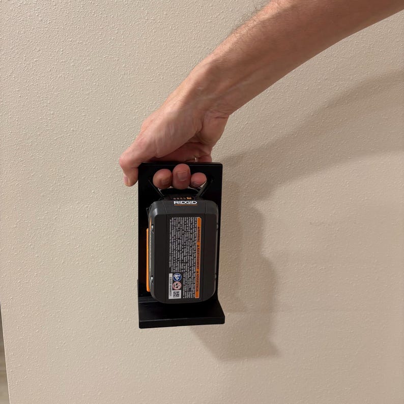 May include: Black metal battery holder with a black and orange Ridgid battery. The holder is designed to mount on a wall. The battery has a handle and a warning label.