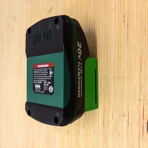 May include: A black and green 20V FLEXPOWER Lithium-Ion battery. The battery has a green center section with a warning label and a black top and bottom. The battery is mounted on a wooden surface.