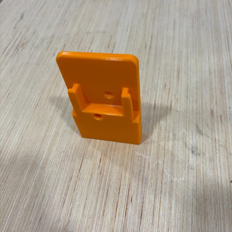 May include: An orange, 3D-printed object with a rectangular back and two supporting arms. A small hole is visible on the front. The object is set against a light wood grain surface.
