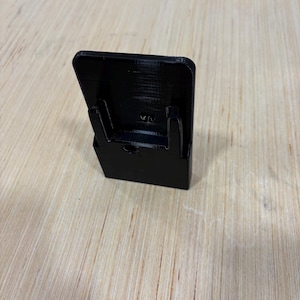 May include: A black 3D-printed phone stand. The stand has a rectangular back with two supporting arms and a base. The stand is designed to hold a phone upright. The stand is made of black plastic.