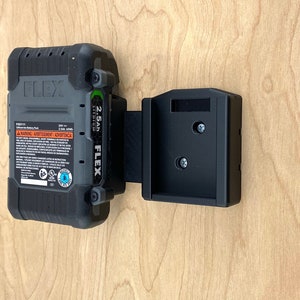 May include: A black and gray FLEX 2.5Ah lithium-ion battery pack mounted on a black holder. The battery pack has the FLEX logo and warning labels. The holder is attached to a light wood surface.