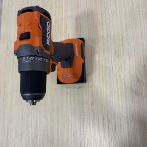 May include: An orange and black cordless drill with the brand name "RIDGID" printed on the side. The drill has a black chuck and a gray adjustable collar. The drill is mounted on a wooden surface.