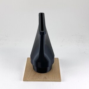 Puede incluir: Una boquilla negra impresa en 3D con una abertura rectangular en la parte superior, sentada sobre un trozo de madera cuadrado.