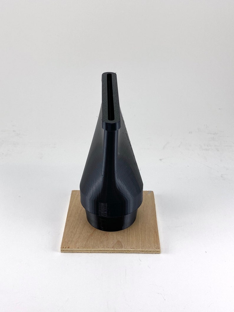 Puede incluir: Una boquilla negra impresa en 3D con una abertura rectangular en la parte superior, sentada sobre un trozo de madera cuadrado.