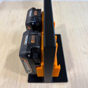 May include: A black and orange battery holder with two black and orange Ridgid lithium-ion batteries. The holder is made of plastic and has a rectangular base. The batteries are labeled with the Ridgid brand name.