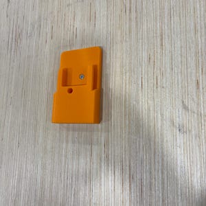 May include: An orange plastic bracket with a rectangular base and a raised section with a screw. The bracket has two side supports and a circular hole. The item is on a light wood surface.