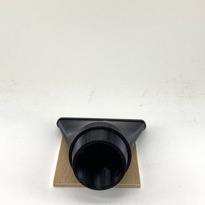 Puede incluir: Boquilla de recolecci&oacute;n de polvo de pl&aacute;stico negro con una abertura redonda. La boquilla est&aacute; sentada sobre un trozo de madera.
