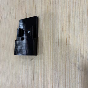 May include: A black, rectangular 3D-printed object with a bracket and a screw. The object has a textured surface and is positioned on a light-colored wooden surface. The object appears to be a mounting bracket.