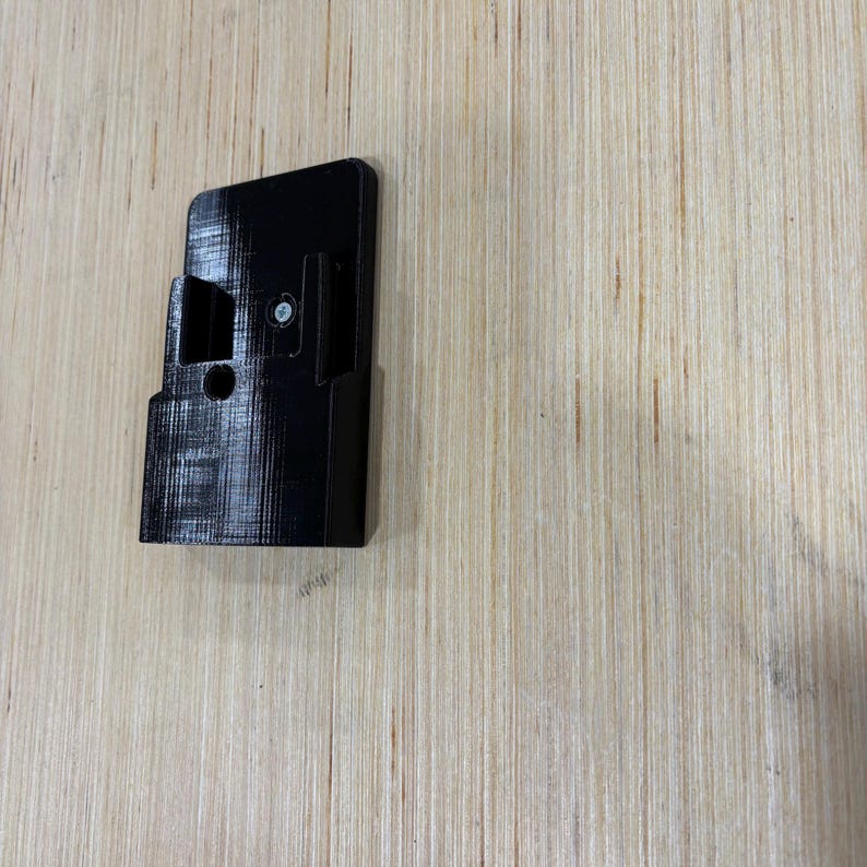 May include: A black, rectangular 3D-printed object with a bracket and a screw. The object has a textured surface and is positioned on a light-colored wooden surface. The object appears to be a mounting bracket.