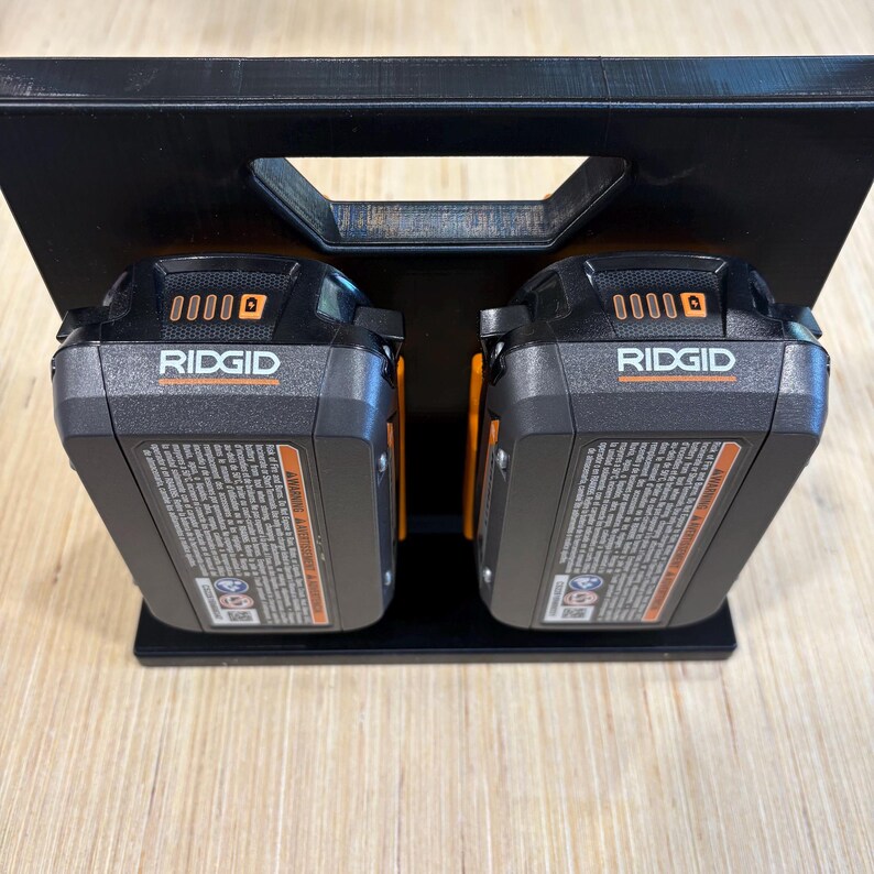 May include: Black power tool battery holder with two black and orange RIDGID batteries. The batteries are in a black holder with a handle. The batteries have the RIDGID logo and other text on them. The holder is on a light brown surface.