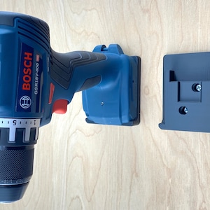 May include: A blue and black Bosch GSR18V-400 drill with a matching wall mount. The drill is mounted on a light wood surface. The drill has a black chuck and a red switch.