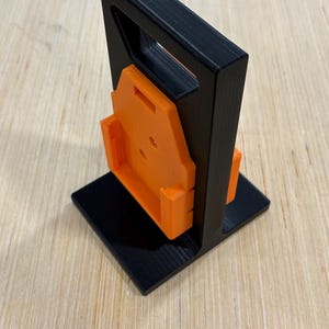 May include: A black and orange 3D-printed tool holder. The holder has a square base, a vertical frame with a handle, and an orange bracket for holding a tool. The design is modern and functional.