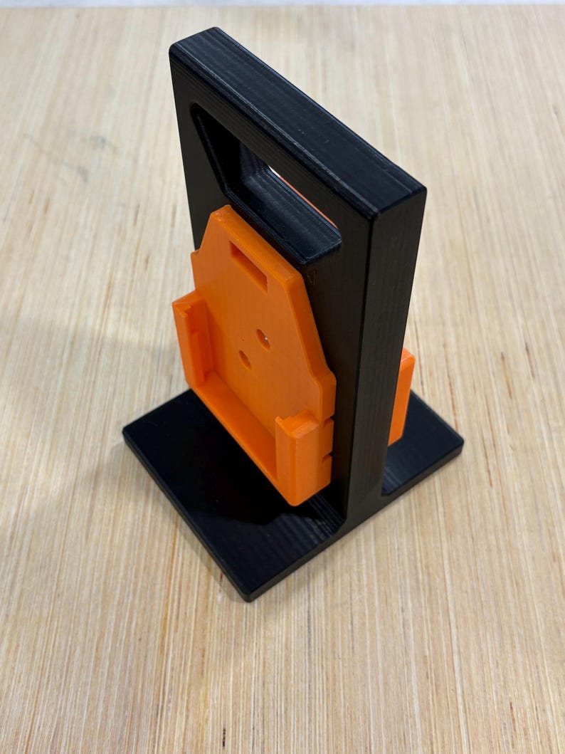 May include: A black and orange 3D-printed tool holder. The holder has a square base, a vertical frame with a handle, and an orange bracket for holding a tool. The design is modern and functional.