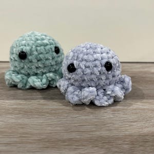 May include: Two crocheted octopus plushies, one in a light blue color and the other in a light green color. Both plushies have black eyes.