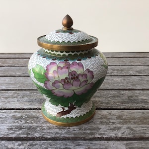 May include: A white cloisonné porcelain jar with a floral design. The jar has a lid with a gold knob and is decorated with green, pink, and white flowers.