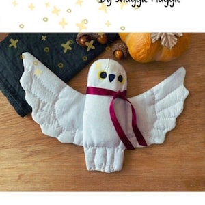 May include: A white plush Hedwig owl with a burgundy ribbon around its neck. The owl is flying with its wings spread out. The image includes the text "Hedwig plush pattern" and "By Snuggle Muggle".