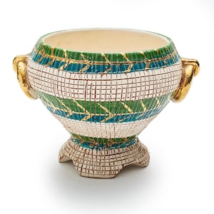 May include: A decorative ceramic vessel with a cream-colored interior and exterior. The exterior features a grid pattern with green, teal, and gold accents. It has two gold handles and a pedestal base.