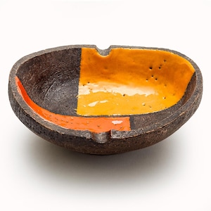 May include: A handcrafted ceramic ashtray with a textured, dark brown exterior. The interior features a geometric design with sections of orange and yellow glaze. The ashtray has a small notch for resting cigarettes.