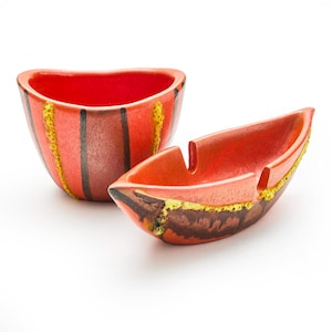 May include: Two ceramic ashtrays. One is a small bowl, the other is boat-shaped. Both are red with black and yellow stripes and accents. The ashtrays are on a white background.