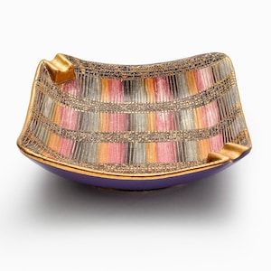 May include: A retro ashtray with a distinctive design. It has a purple base and gold-coloured trim. The interior features a colourful pattern of vertical stripes in pink, orange, grey, and gold. The ashtray has two built-in cigarette rests.