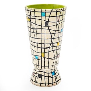 May include: A tall, cream-colored ceramic vase with a green interior. The vase features a black grid pattern with colorful square accents. The base has a flared design, and the vase has a mid-century modern aesthetic.