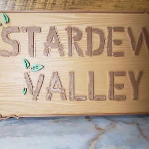 May include: A wooden sign with the words "Stardew Valley" in brown raised letters. The sign has a rustic wood grain texture and is decorated with small green leaves.