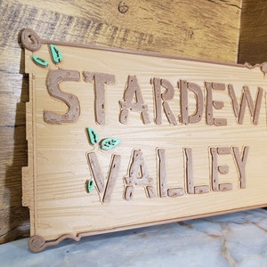 May include: A wooden sign with the words "Stardew Valley" in brown raised letters. The sign has a light brown wood grain texture and is decorated with green leaves.