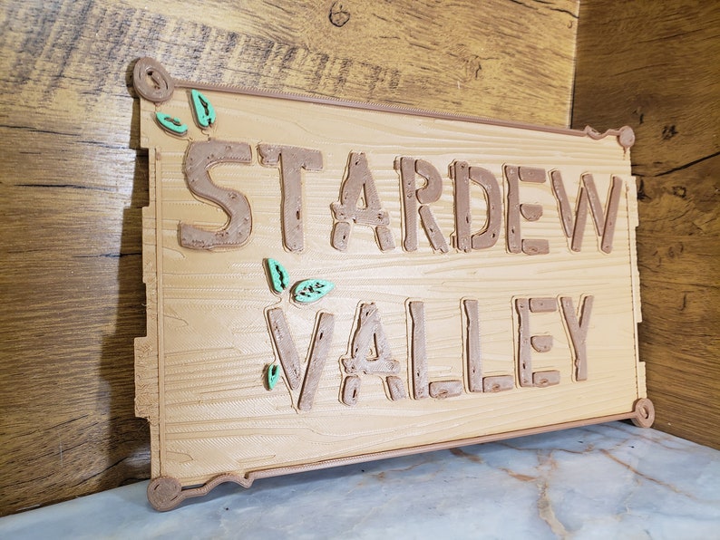 May include: A wooden sign with the words "Stardew Valley" in brown raised letters. The sign has a light brown wood grain texture and is decorated with green leaves.