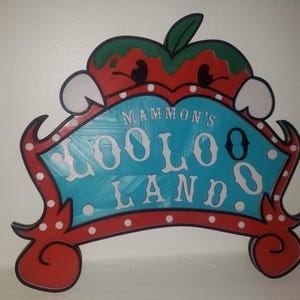 May include: A red and white sign with a blue background and white polka dots. The sign says "Mammon's LooLoo Land" in white letters. The sign is shaped like a cartoon apple with a green stem and red and white details.