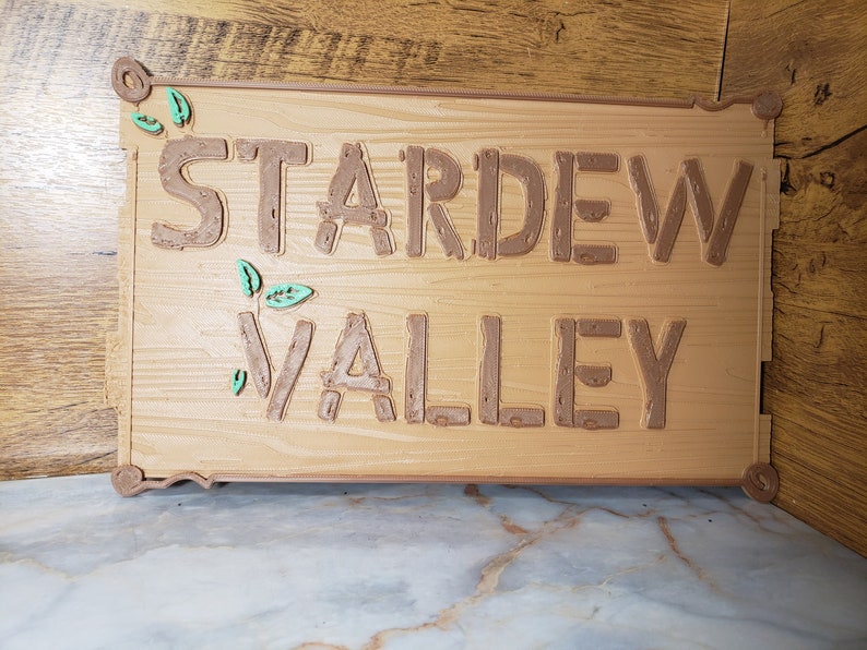 May include: A wooden sign with the words "Stardew Valley" in brown raised letters. The sign has a rustic wood grain texture and is decorated with small green leaves.