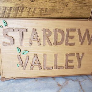 May include: A wooden sign with the words "Stardew Valley" in brown raised letters. The sign has a brown background and green leaves on either side of the text.