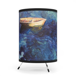 May include: A table lamp with a cylindrical lampshade featuring a painting of a small boat on a blue and green water surface. The lamp has a black base and three silver legs.
