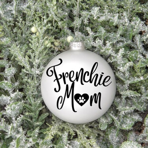 May include: White Christmas ornament with the text "Frenchie Mom" in black cursive lettering and a black paw print inside a heart.