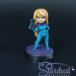 Puede incluir: Una figura miniatura pintada de un personaje femenino con un traje espacial azul y morado que sostiene una pistola. La figura está de pie sobre una base morada y negra.