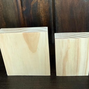 May include: Two unfinished wooden blocks. One is rectangular with a slanted side, the other a smaller cube. The wood grain is visible, and the blocks are light in colour. They are likely for crafts or display.