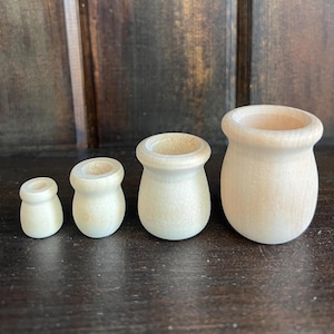 May include: Four small, unfinished wooden pots of varying sizes are arranged in a row. The pots have a light, natural wood color and a simple, rounded design. The background is a dark wooden surface.