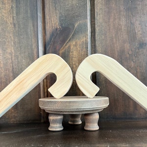 May include: Two unfinished wooden candy cane shapes are propped up against each other on a small wooden pedestal with three legs. The candy canes are light brown and the pedestal is dark brown.