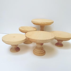 May include: Five unfinished wooden pedestal cake stands with round tops and decorative bases. The stands vary in height and diameter, showcasing natural wood grain. Perfect for displaying desserts or decorative items.