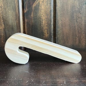 May include: A wooden candy cane shape, unpainted, with a curved hook and a long, straight body. The wood grain is visible, with alternating light and dark stripes. The candy cane is resting on a dark wooden surface.