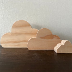May include: Three wooden cloud shapes in different sizes, arranged from largest to smallest. The clouds are made of light brown wood and have a smooth, unfinished surface.