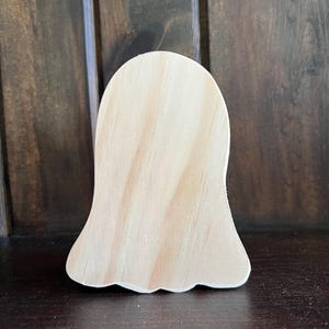 May include: A light-colored, unfinished wooden ghost-shaped cutout. The cutout has a rounded top and a scalloped bottom edge. The wood grain is visible, and the cutout is set against a dark brown background.