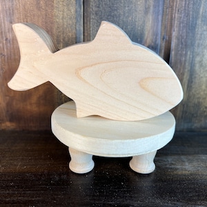 May include: A wooden fish figurine on a small wooden stand with three legs. The fish is light brown and has a simple design. The stand is also light brown and has a smooth surface.