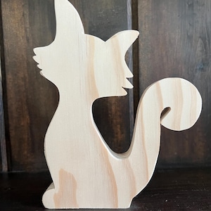 May include: A wooden cat figurine, cut from light-colored wood, stands upright. The cat's silhouette features a curved tail and pointed ears. The wood grain is visible, adding texture to the simple design.