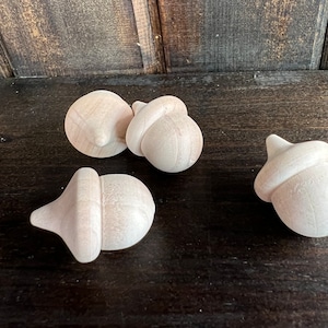 May include: Four unfinished wooden acorn ornaments. The acorns are a light brown color and have a smooth, rounded surface.