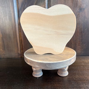 May include: A wooden apple shape sits on a small wooden pedestal with three legs. The apple is unfinished and ready to be painted or decorated.