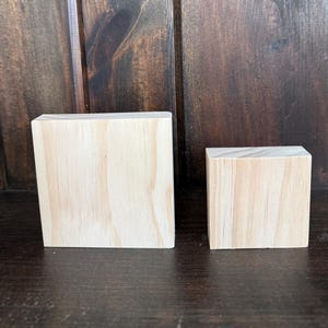 May include: Two unfinished wooden blocks of different sizes. The larger block is square, and the smaller block is a cube. The wood grain is visible, and the blocks are set against a dark wood background.