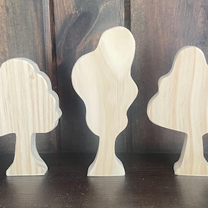 May include: Three wooden tree figurines with natural wood grain. The trees have different shapes, with rounded and abstract designs. The background is a dark brown wooden surface.