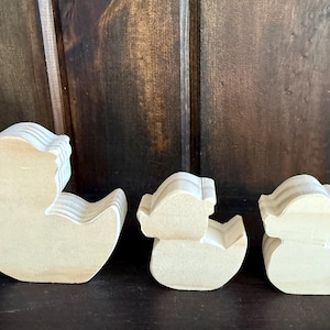 May include: Three wooden duck cutouts of varying sizes. The ducks are light-colored and appear to be unfinished, with a layered design. They are arranged in a row against a dark wooden background.