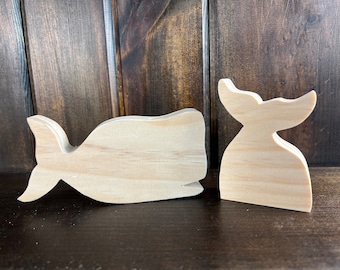 May include: Two unfinished wooden whale shapes. The larger whale is laying down with its tail facing the left side of the image. The smaller whale is standing upright with its tail facing the right side of the image.